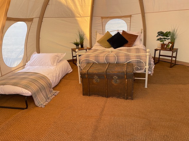 Inside a CampAble tent: a double bed with cushions and a tartan throw, a single bed, bedside tables and plants